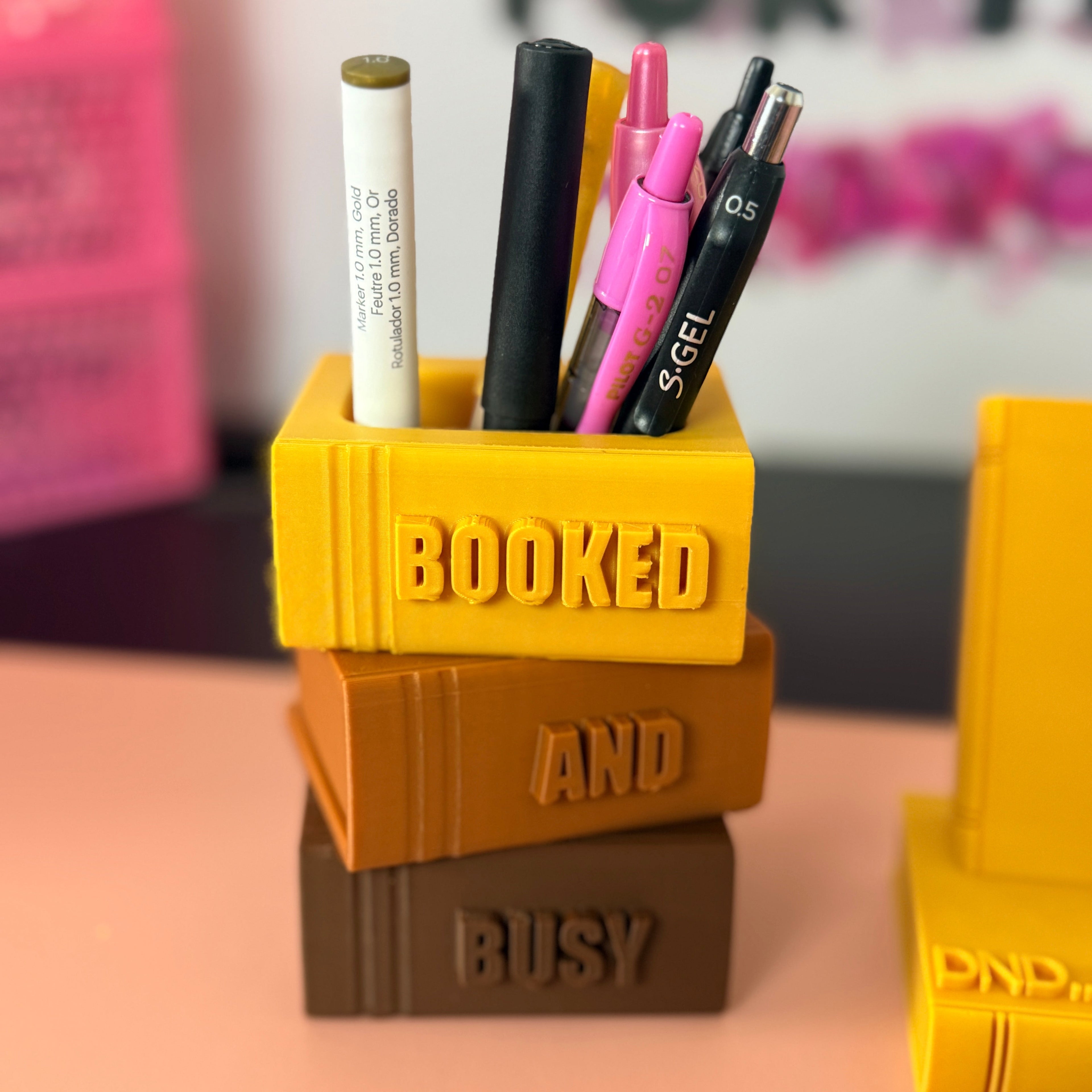 Yellow, brown, and orange book-shaped pen holder with stationery items on a pink surface.